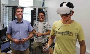 A professor and a student look on as another student wears a virtual reality headset.