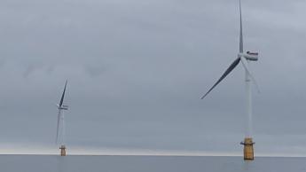 Two wind turbines rise from the surface of the ocean in front of an overcast sky