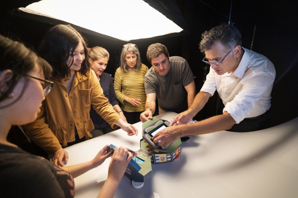 Four students and two professors gather around a lit desk to tinker with a cardboard package decorated with Pokemon characters.