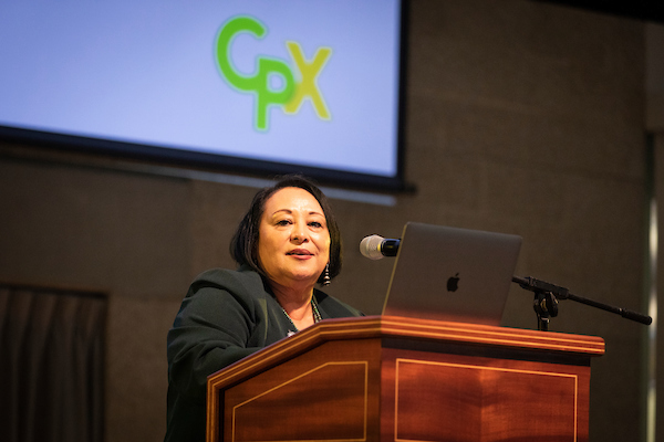 Vice President Jozi de Leon speaks from a podium, in front of a screen displaying the letters CPX