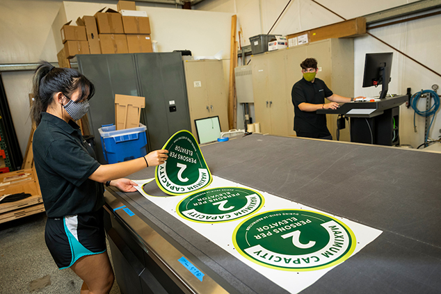 Michaela Kwan, left, and Chris Jones work with the die cut machine at the Cal Poly Printing Press as they and other student employees print signage to be used in buildings on campus during the  coronavirus pandemic. 