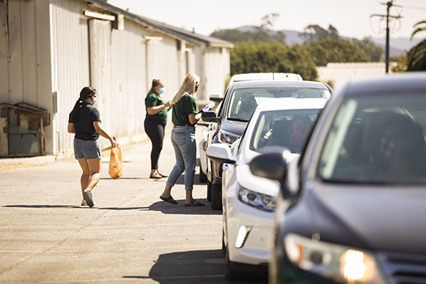 Three cars wait in line for ice cream while three Cal Poly students carry bags with orders out to the cars.