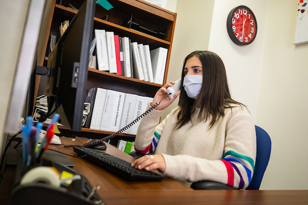 A student wearing a light blue mask and a white striped sweater answers a desk phone at the Low Income Taxpayer Clinic.