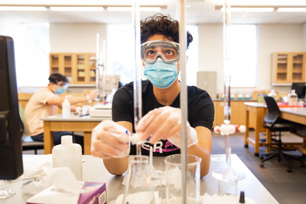 A student wearing a surgical mask and chemistry goggles performs an acid titration procedure in a lab. 