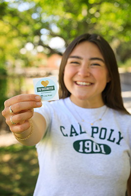 A student wearing a Cal Poly shirt holds out an "I'm vaccinated" sticker.