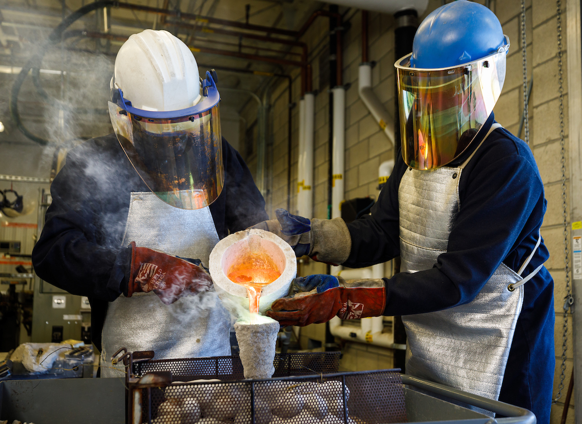 Two students wearing protective gear pour molten metal into a mold.
