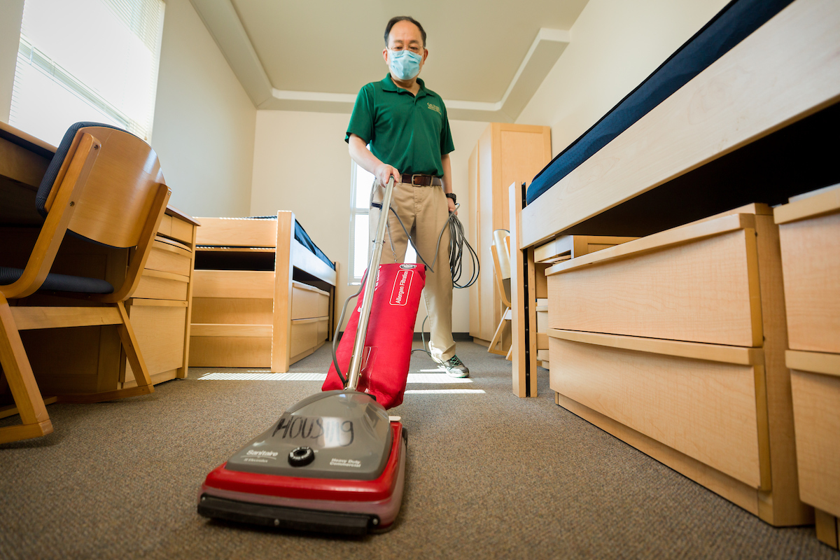 A man wearing a Cal Poly polo shirt and khaki pants wears a mask and vacuums an empty dorm room.
