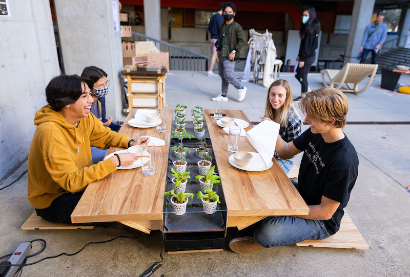 Students sit around a hydroponic table made out of light colored wood and smile.