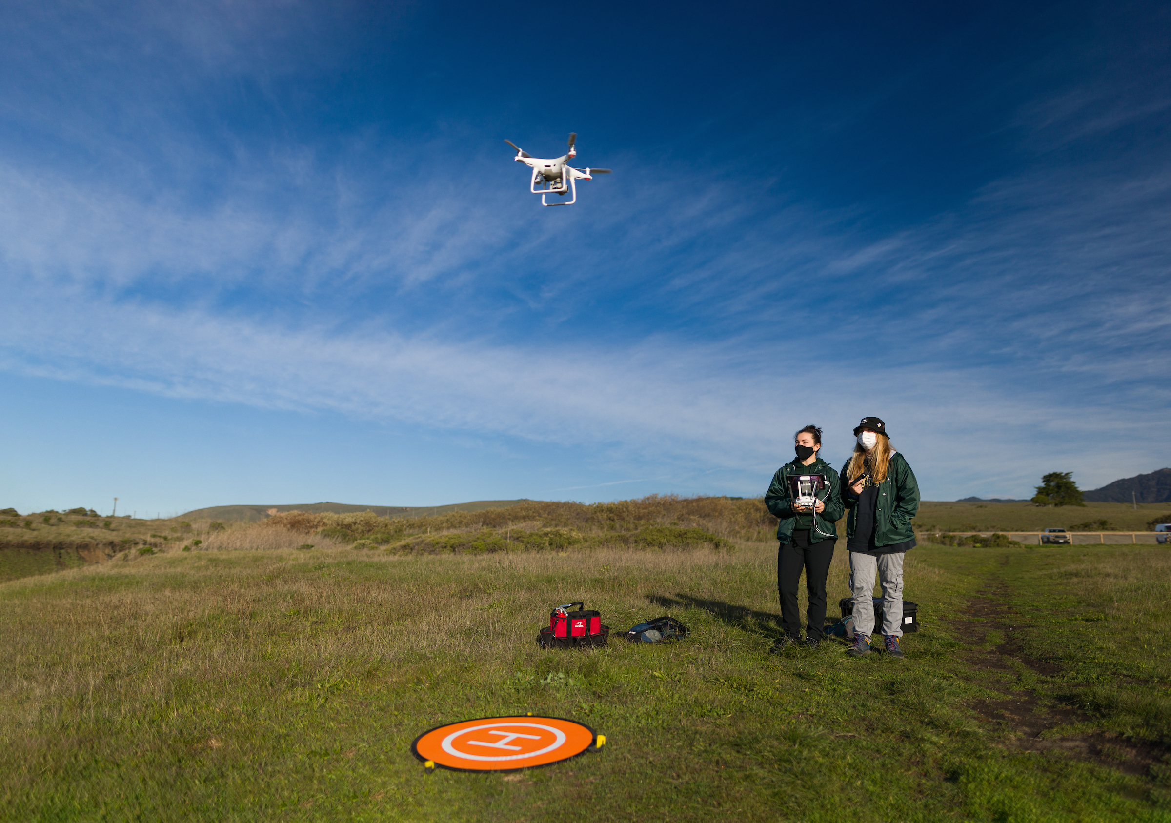 Two women wearing green Cal Poly jackets operate an aerial drone on a grassy bluff. The drone hovers in the foreground.