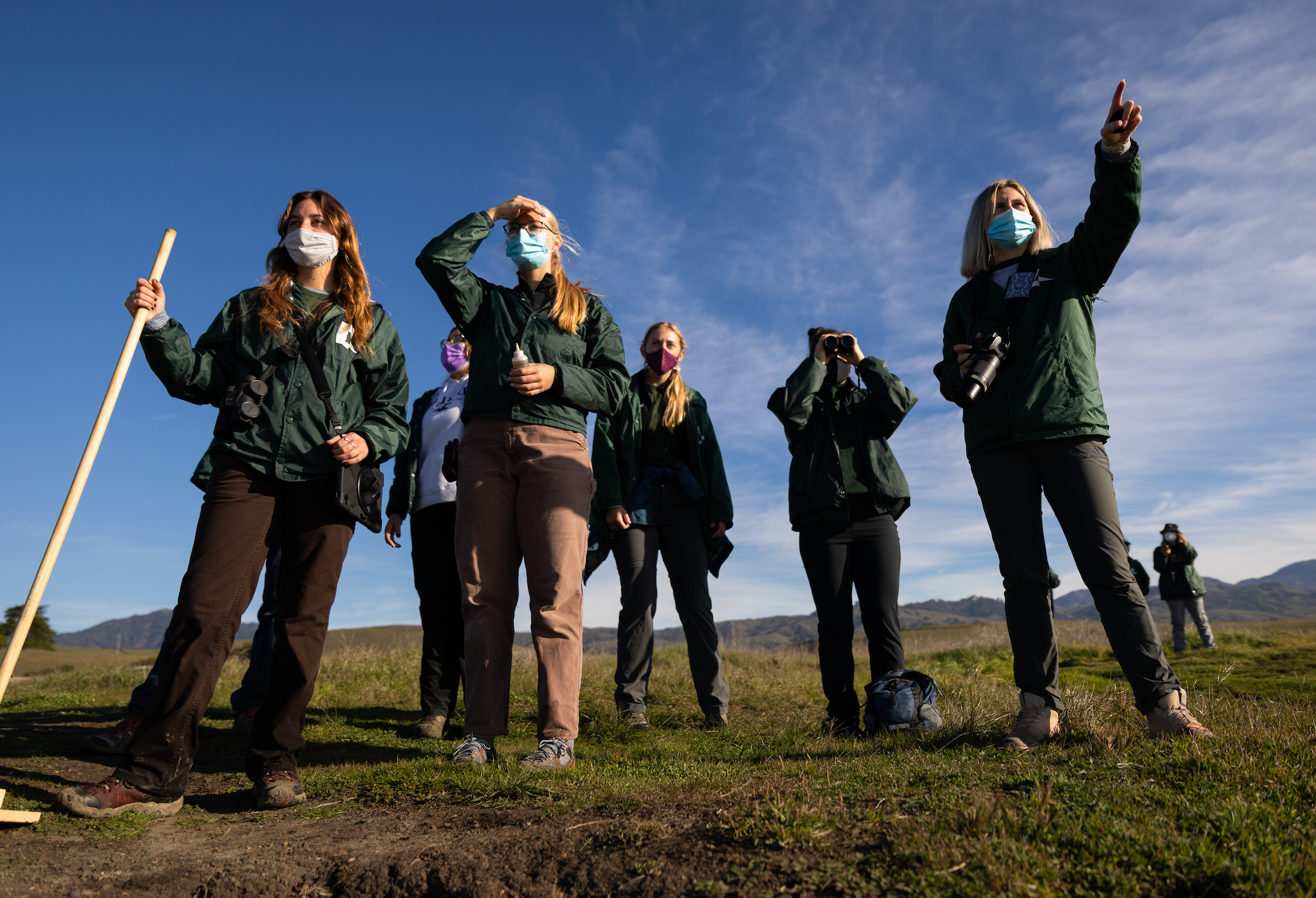 Student researchers wearing green Cal Poly jackets stand on a grassy bluff above a beach with survey equipment. 