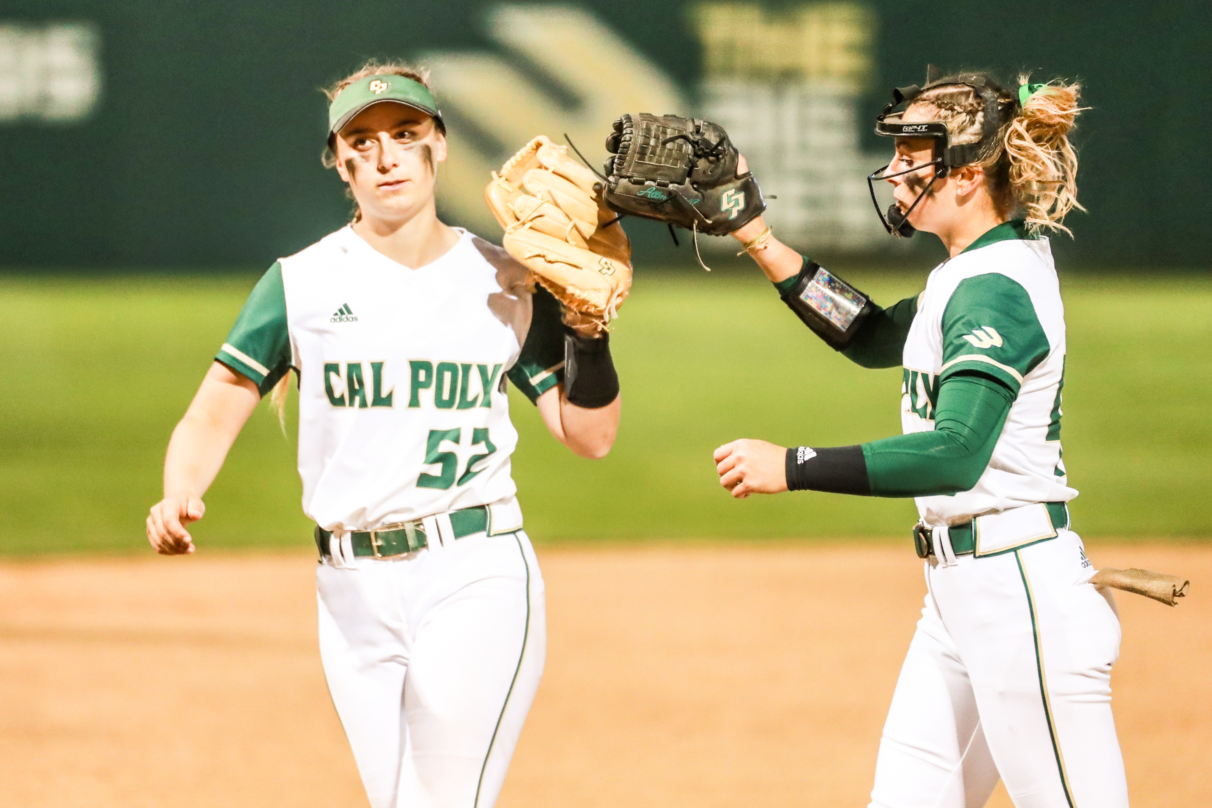 Two sisters wearing Cal Poly softball uniforms high five on the field during a game.