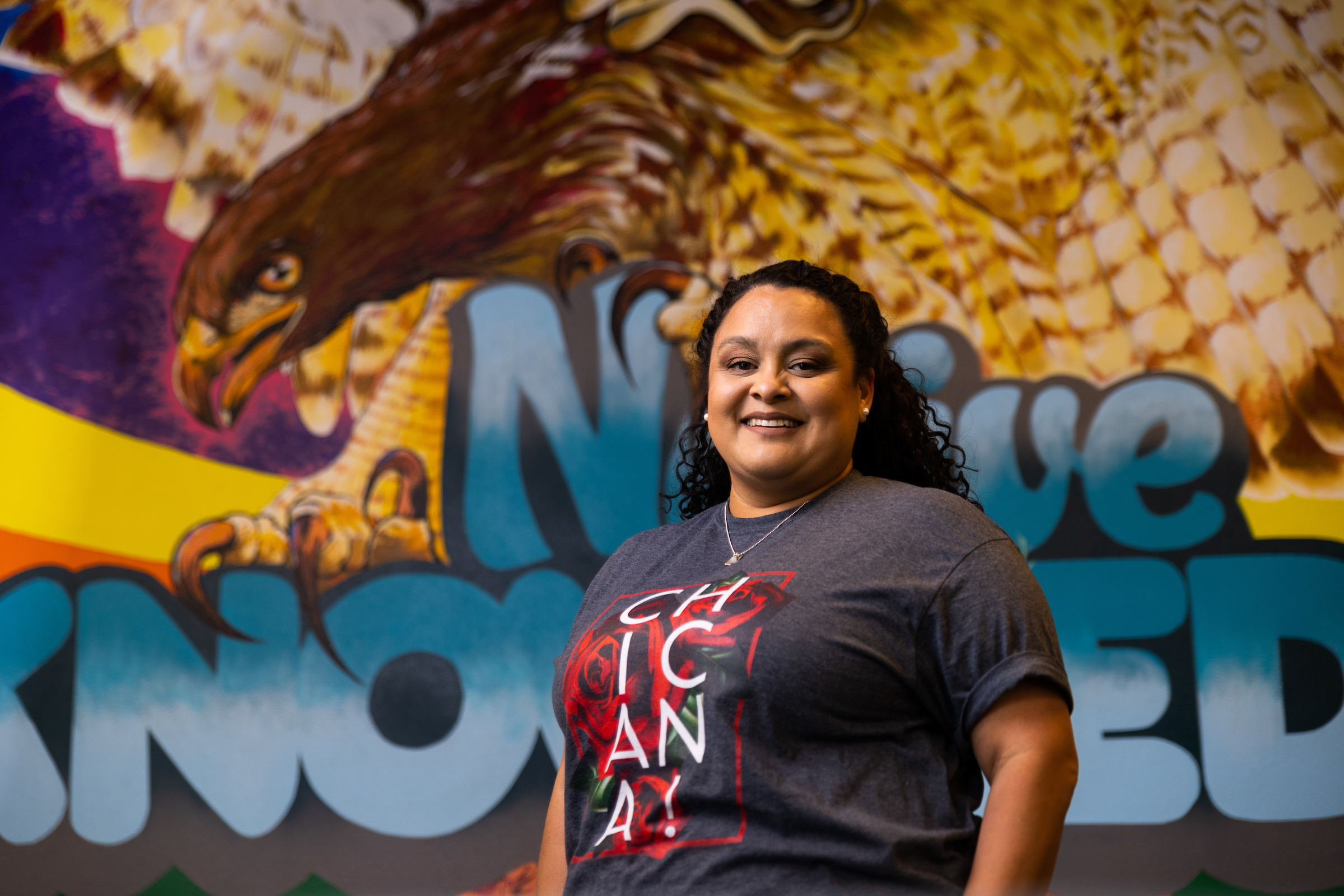 Cheryl Flores smiles in front of the mural at the Native American and Indigenous Cultural Center on campus.