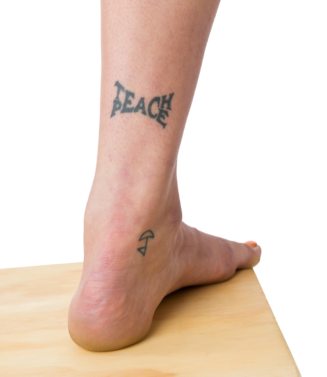 A young woman's ankle, featuring a tattoo that reads "Teach Peace"