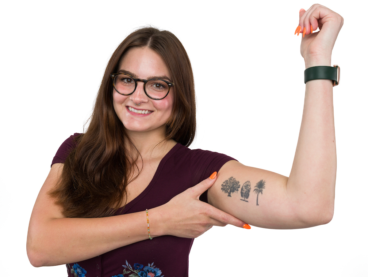 A young woman displays a bicep tattoo featuring three trees
