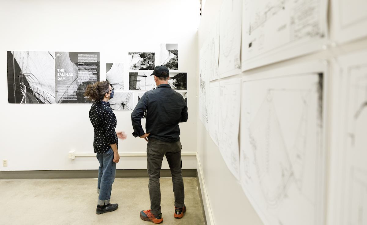 Two people, one wearing a face mask, look at drawings from an architecture studio class regarding their work at the Salinas Dam.