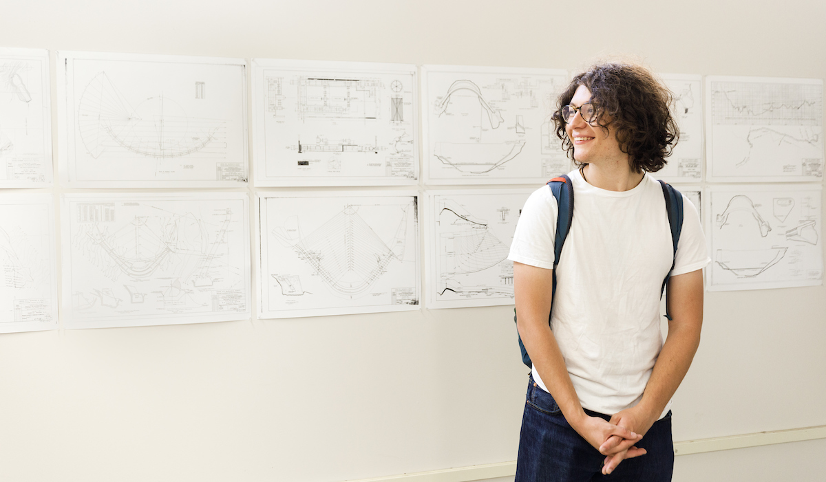 A student stands in front of a wall covered in paper architectural drawings.