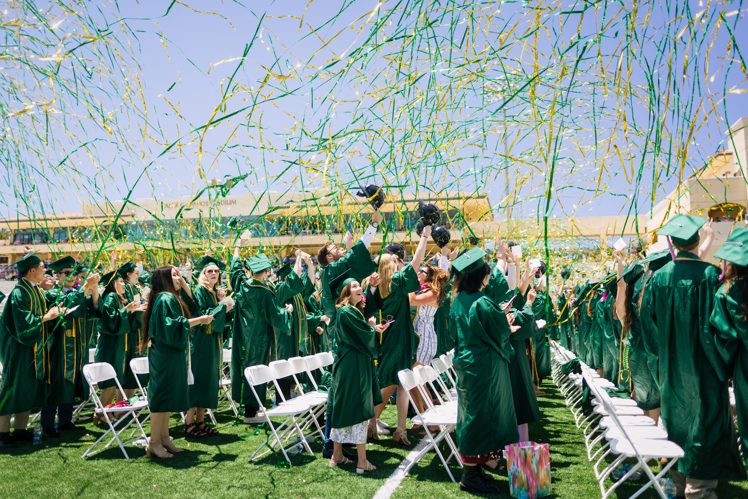 Students in green graduation robes stand at the commencement ceremony while green and gold streamers fly around.
