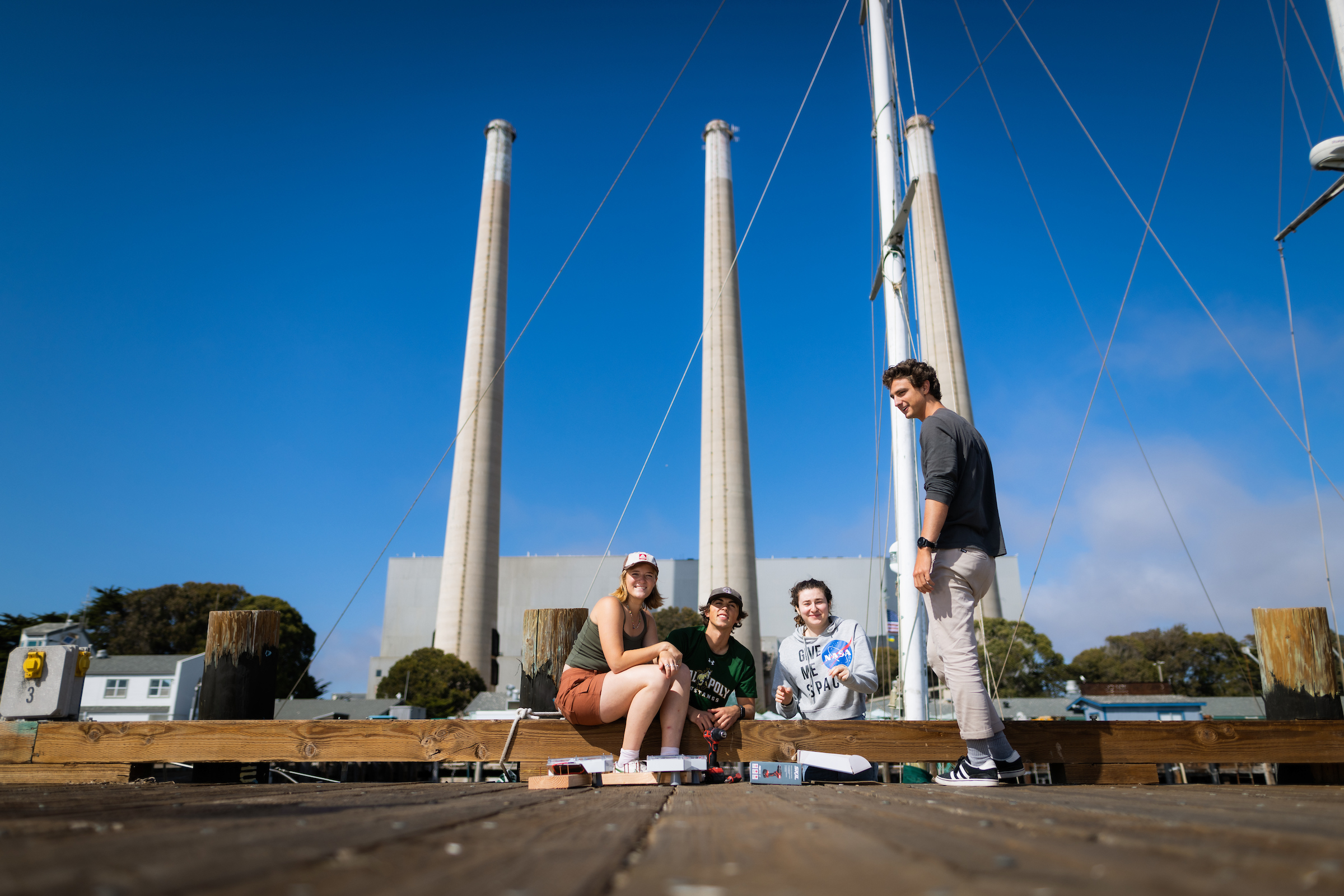 Four students gather on the side of a pier in Morro Bay. Behind them are three smokestacks.