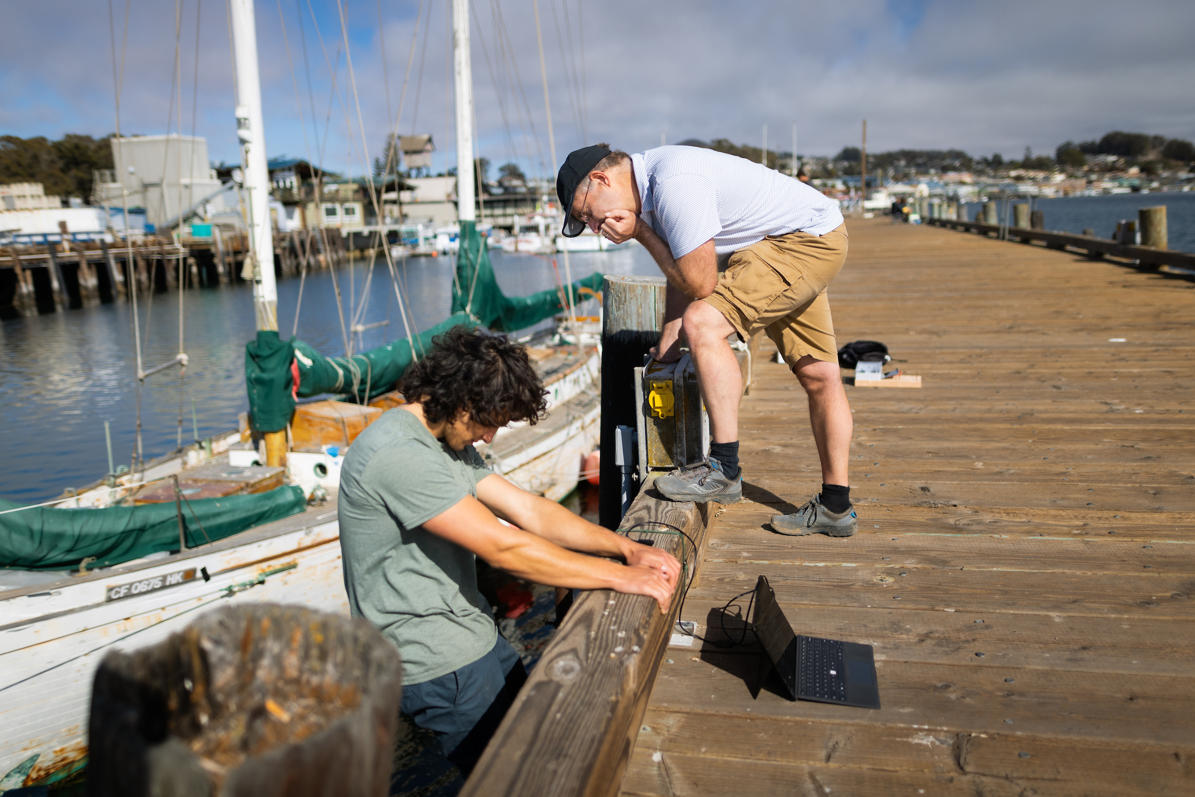 A student hangs off the edge of a pier and his professor leans down to talk to him. They are deploying sensors to measure waves and tides.