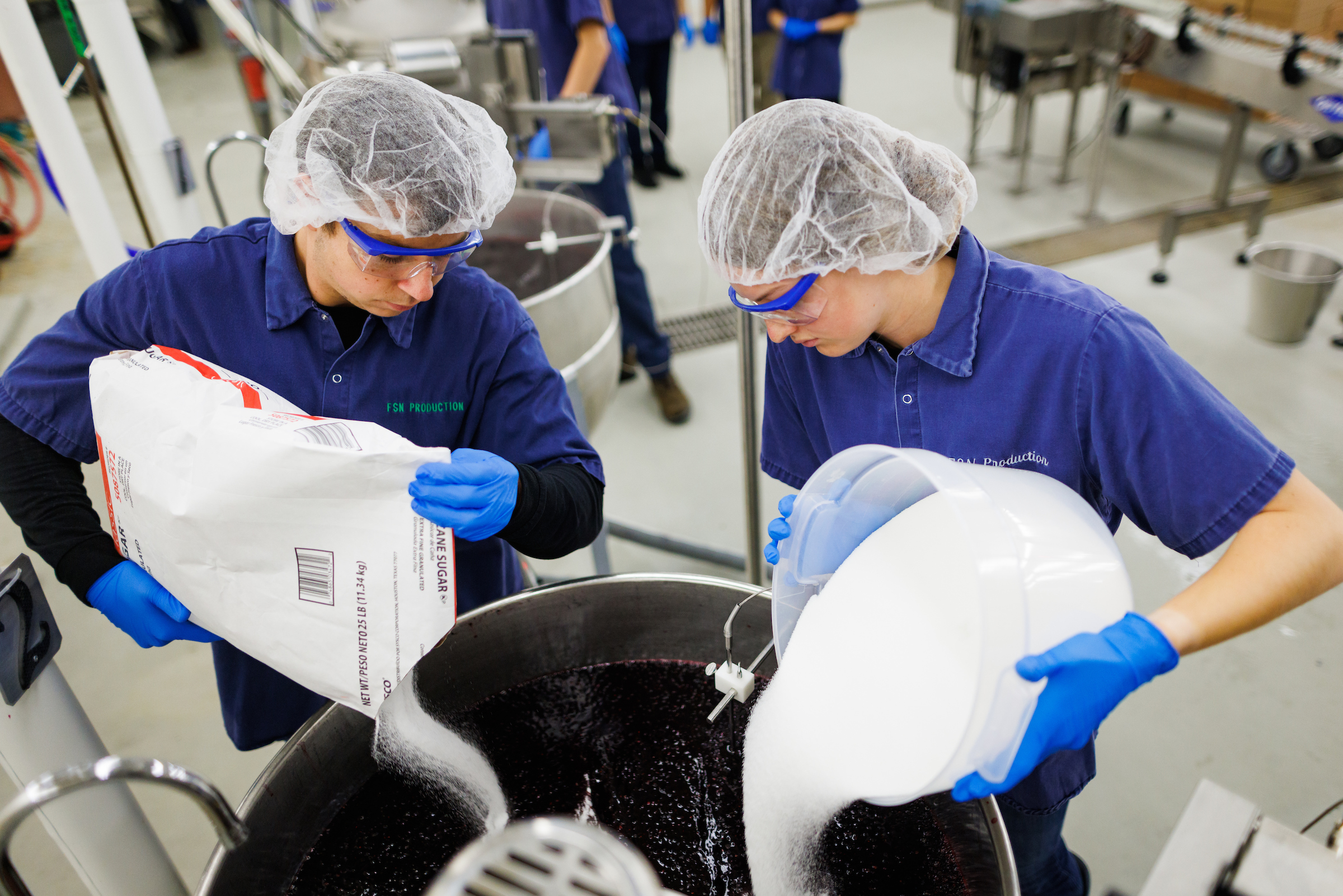 Students wearing safety gear pour sacks of sugar into a batch of jam.