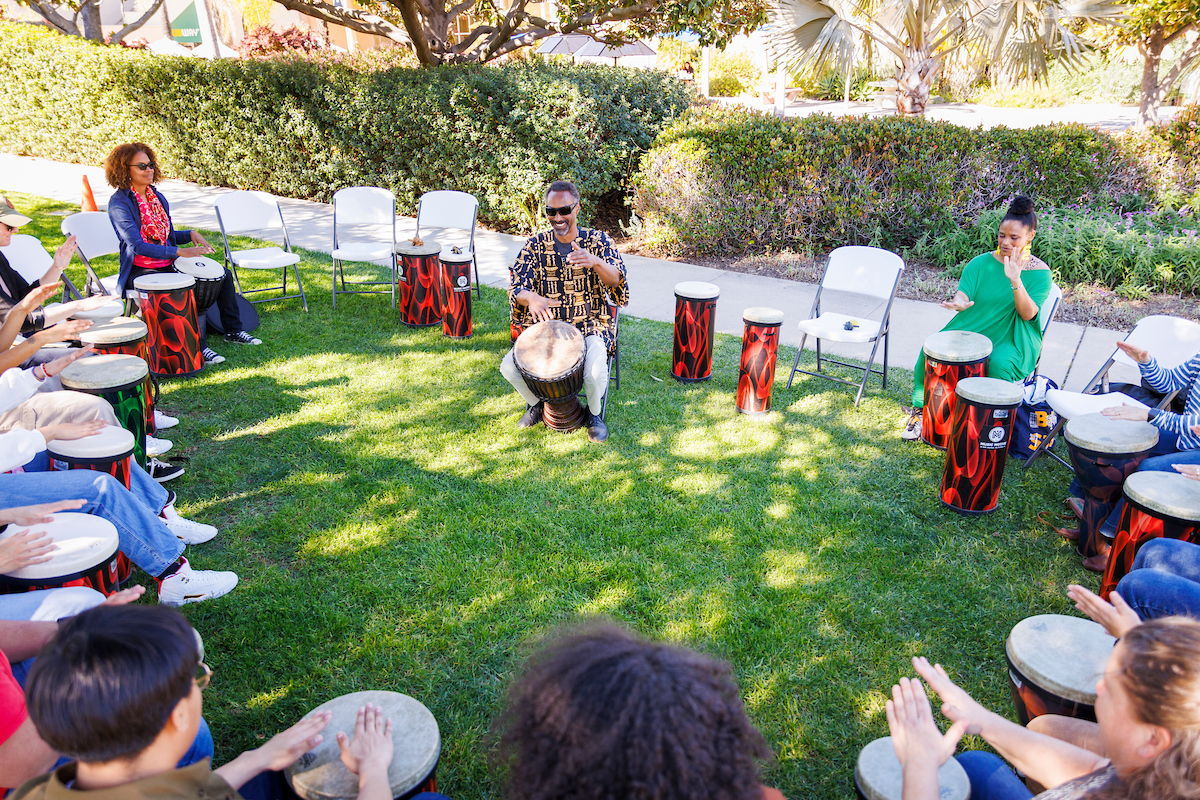 Participants in a drum circle gather around the leader, a Black man in the center, as they learn drumming patterns.