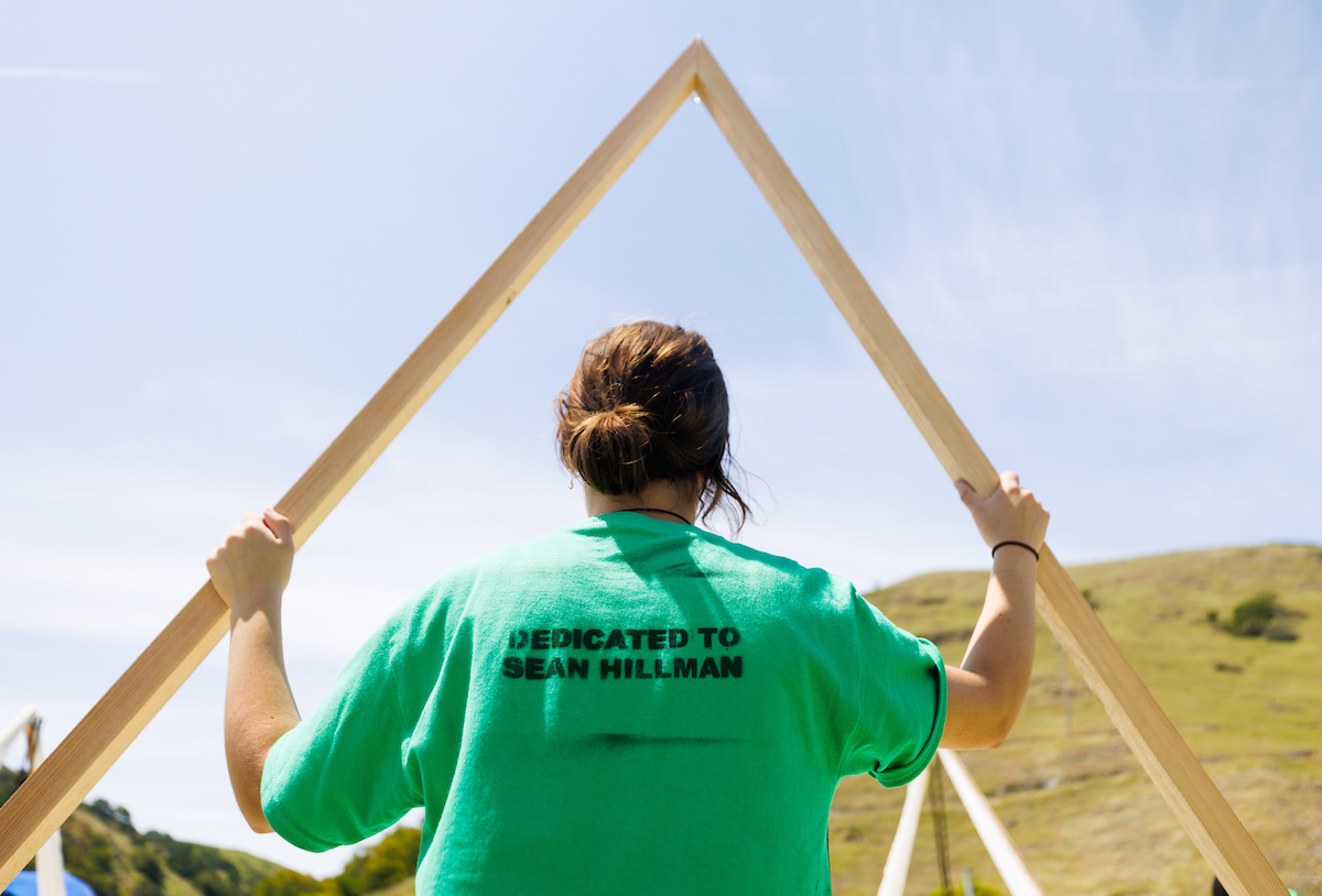 A student wears a green T-shirt that says "Dedicated to Sean Hillman" on the back as they put two wooden beams together. 