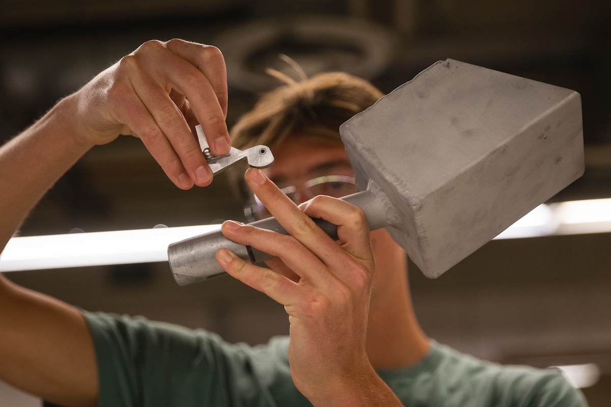 A student wearing safety goggles is in the background and the prototype for a dust tolerant extension handle mechanism, which looks like a small shovel, is the main focus.