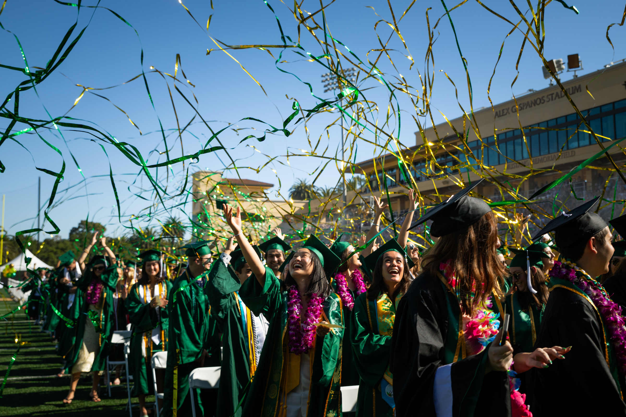 Students in green graduation robes stand at the commencement ceremony and look up smiling while green and gold streamers fly around.