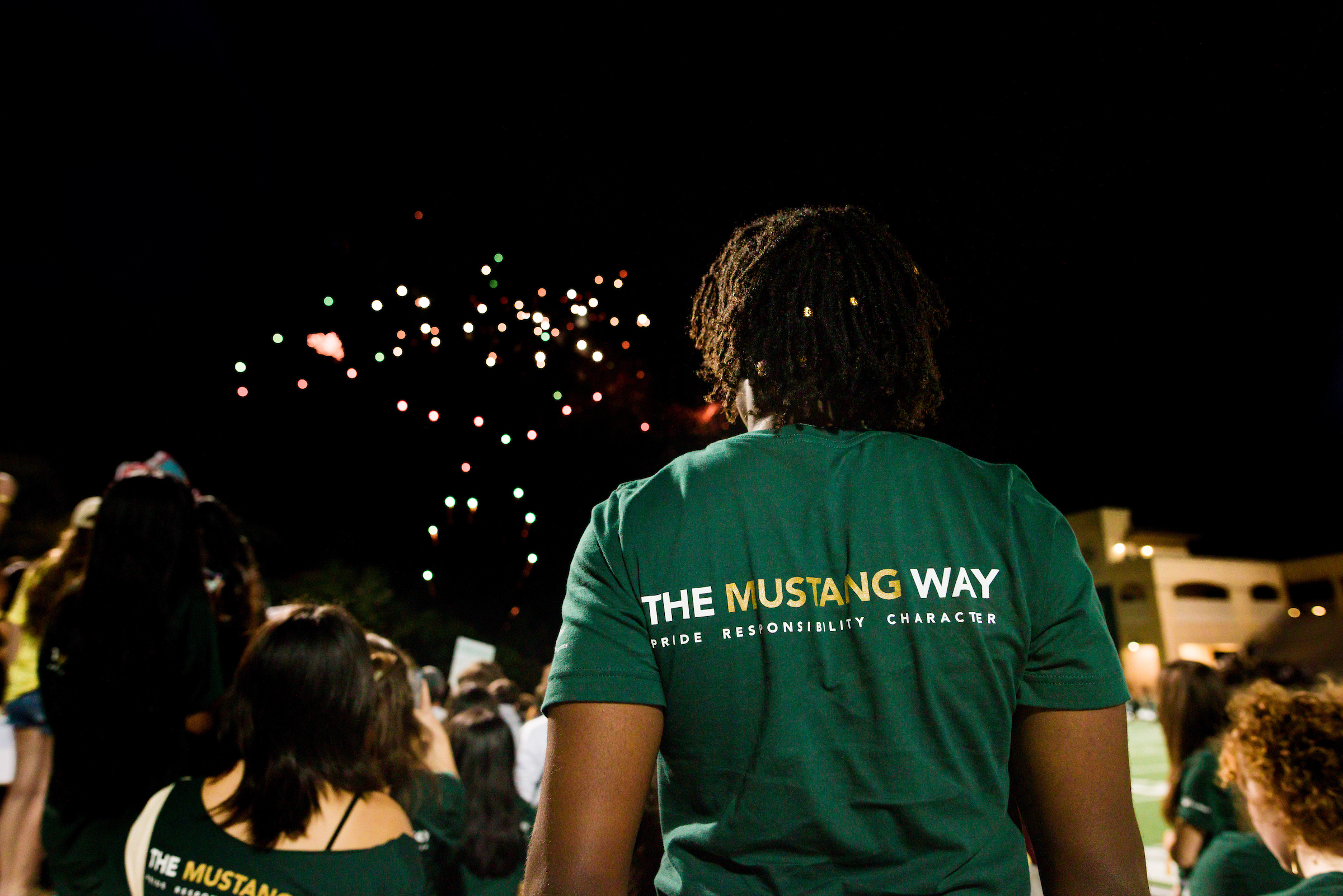 Students look up at a fireworks display above Spanos Stadium. The student in the foreground, whose back is to the camera, is wearing a green shirt that says The Mustang Way across the top.