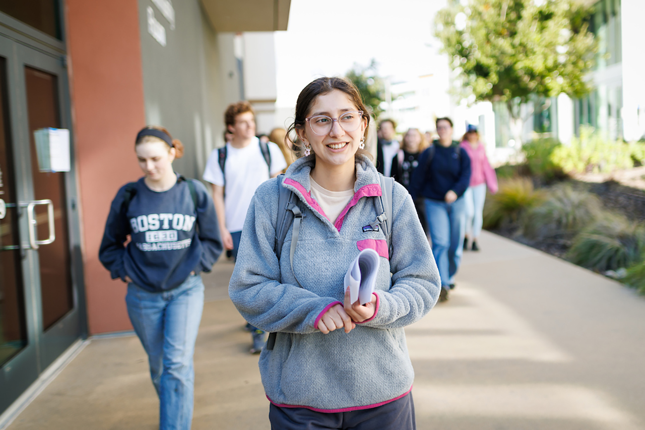 A young woman in a grey sweatshirt and glasses leads a group of students down a campus walkway.