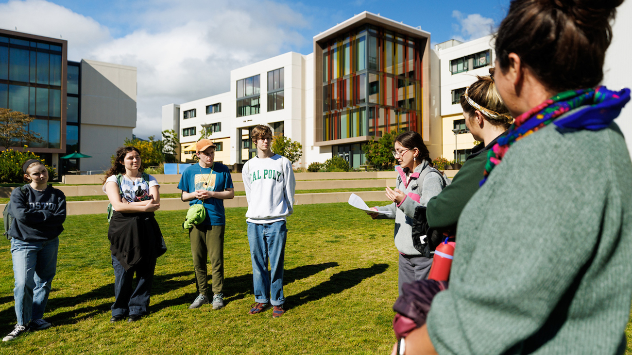 Standing on a lawn in front of a colorful building complex, a young woman in a grey sweatshirt reads from a script as a group of students listens.