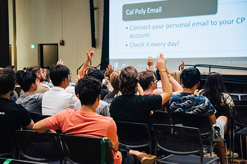 Students raise their hands while seated in a room with information on a screen about checking their Cal Poly email