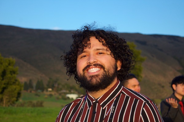 A man with curly black hair and wearing a red, black and white striped shirt smiles at the camera. There are mountains in the background.