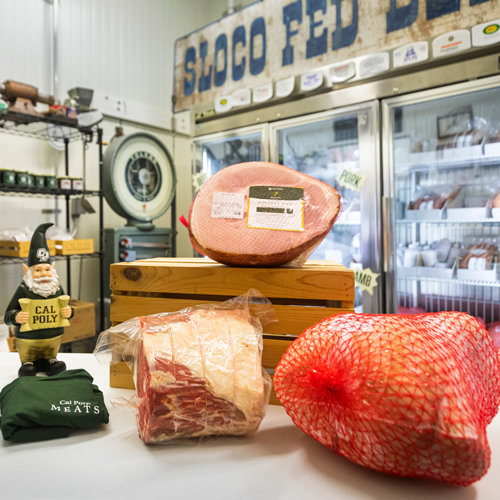 A ham, prime rib and other meat products sit on a table in front of a glass-front refrigerator