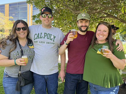 Four people with Cal Poly shirts and hats hold glasses and pose for a photo during at a 2022 alumni wine and beer event at Cal Poly.