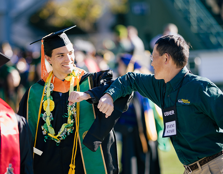 A student bumps elbows with an event staff member at a Fall Commencement ceremony.