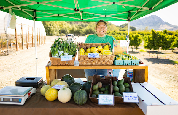 A student smiles behind a booth offering Cal Poly grown produce. 