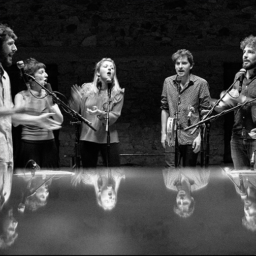 This black and white picture shows singers around a mirrored table, where you can see tiger reflection as they sing into their microphones.