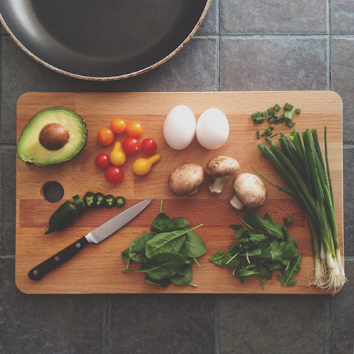 A cutting board with a knife, two eggs and vegetables, including tomatoes, spinach, green onion, avocado and mushrooms.