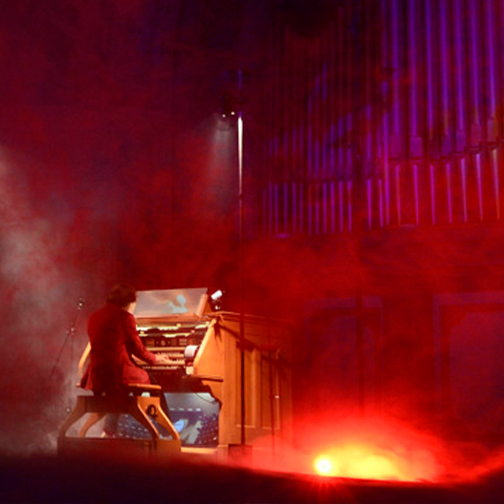 Man in a red coat playing an organ with fog and organ in the background