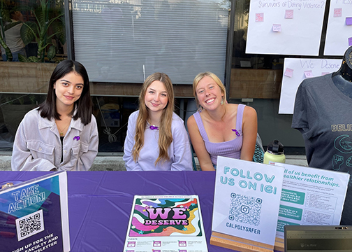 Three female students wearing purple seated at a support table