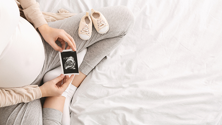 Photo of a pregnant woman, seated with legs crossed, holding an ultrasound photo of her child.