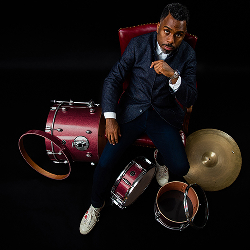  Shot from above, the picture shows Nate looking up at the camera in a suit, sitting in a red chair surrounded by his drum set. 