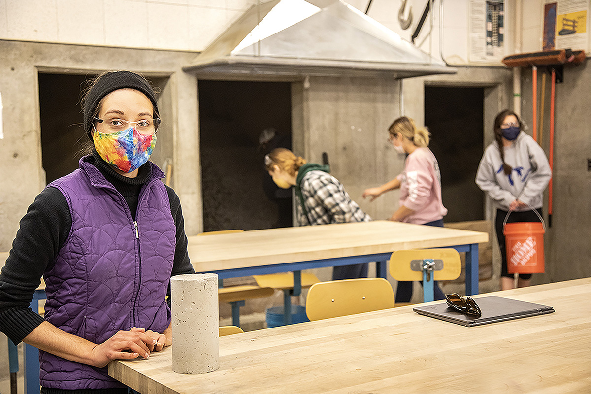 A woman in a purple puffer vest and colorful face mask rests her hands on a wooden table to the left of the photo. In the background are three other students, carrying objects in the lab.