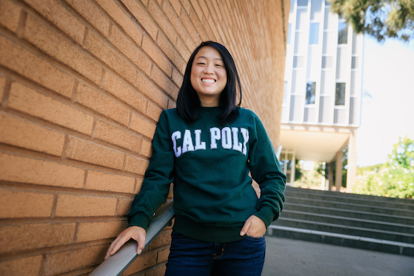 A woman with black hair wearing a green Cal Poly sweatshirt smiles and poses next to a red brick wall.