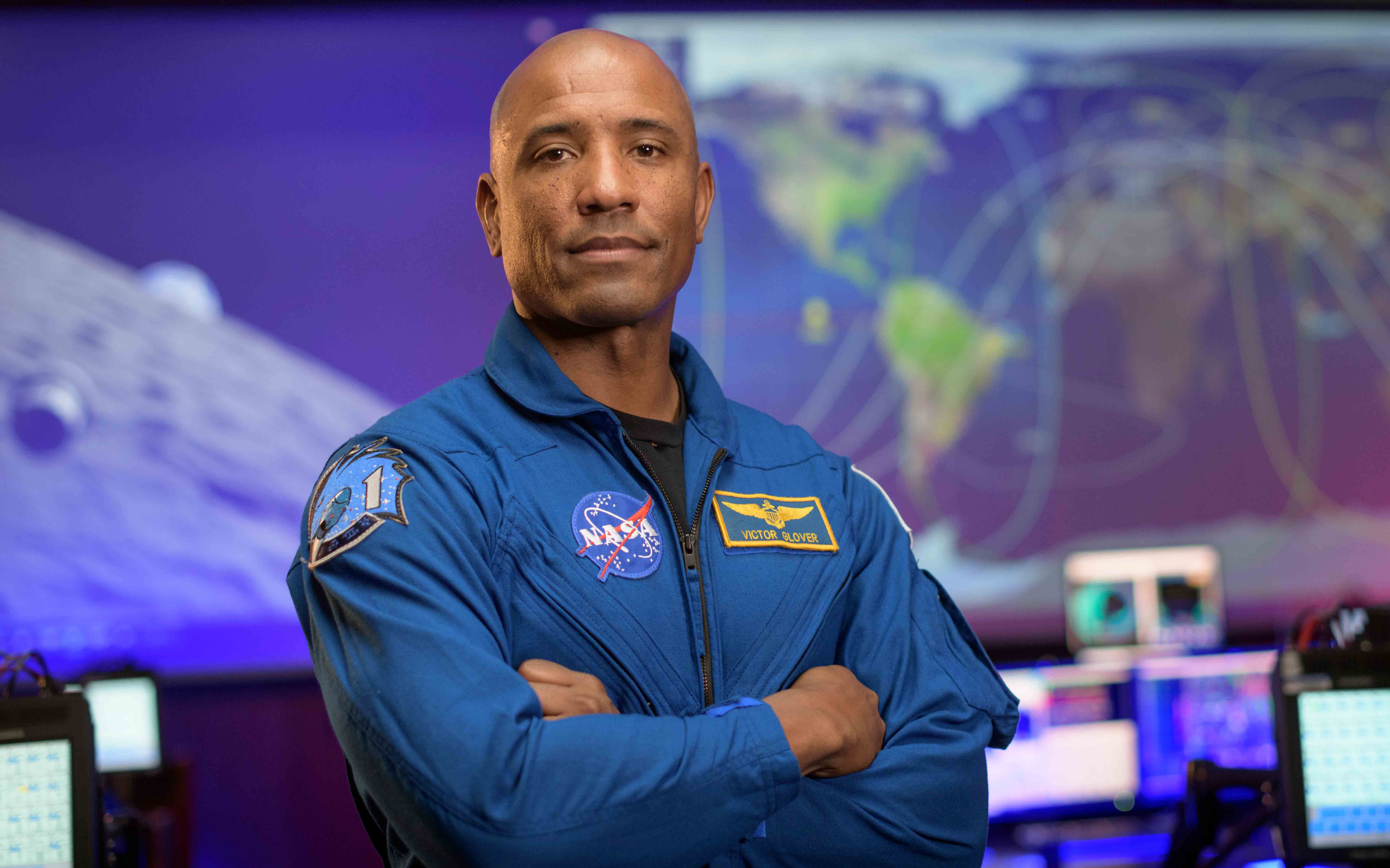 Alumnus Victor Glover wears a blue NASA flight suit and crosses his arms as he poses for a portrait.