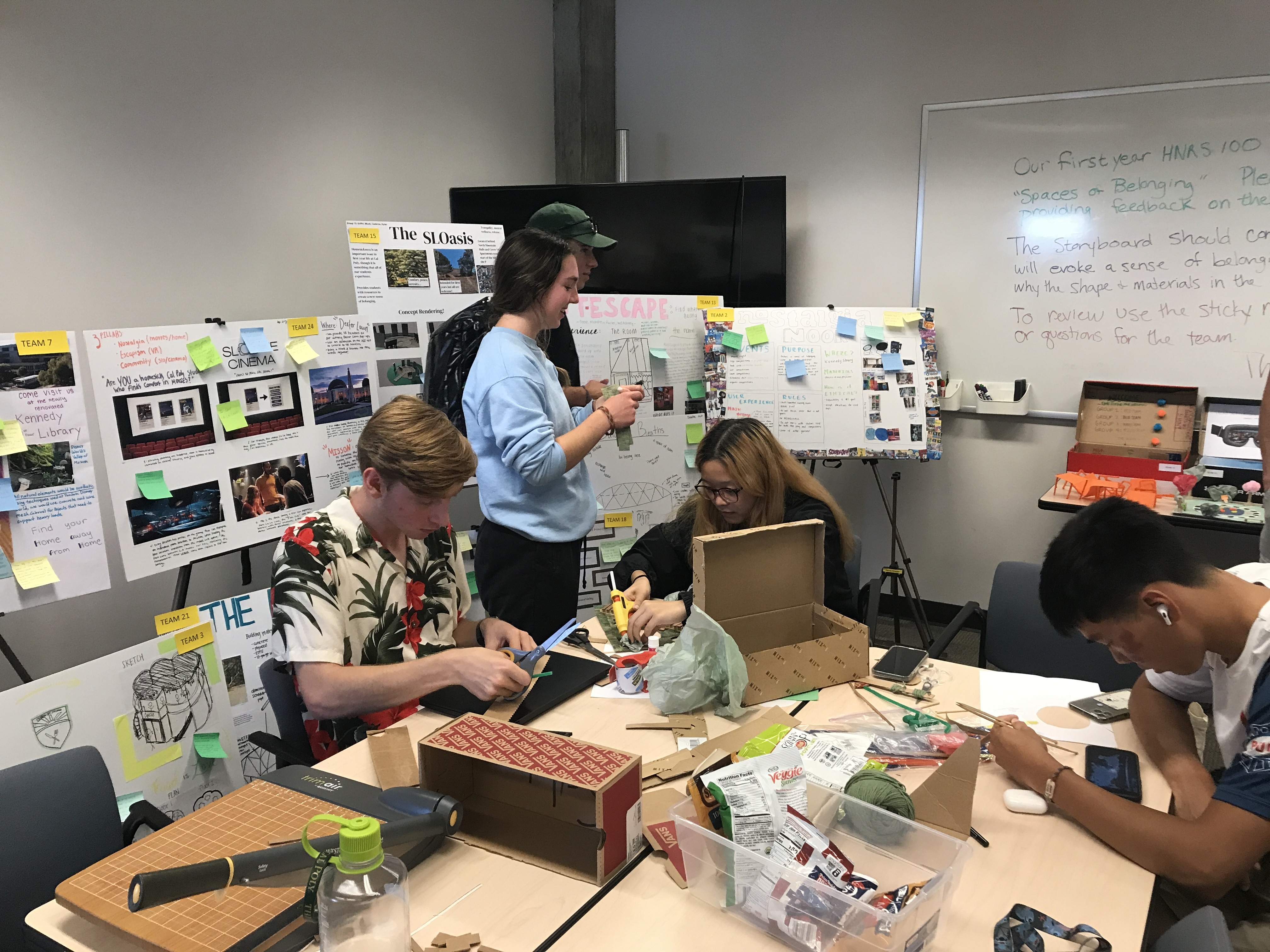 Students gather around a table covered in poster boards and sticky notes as they design a space of belonging.