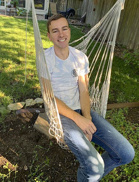A young man in a T-shirt smiles while sitting in a hammock seat