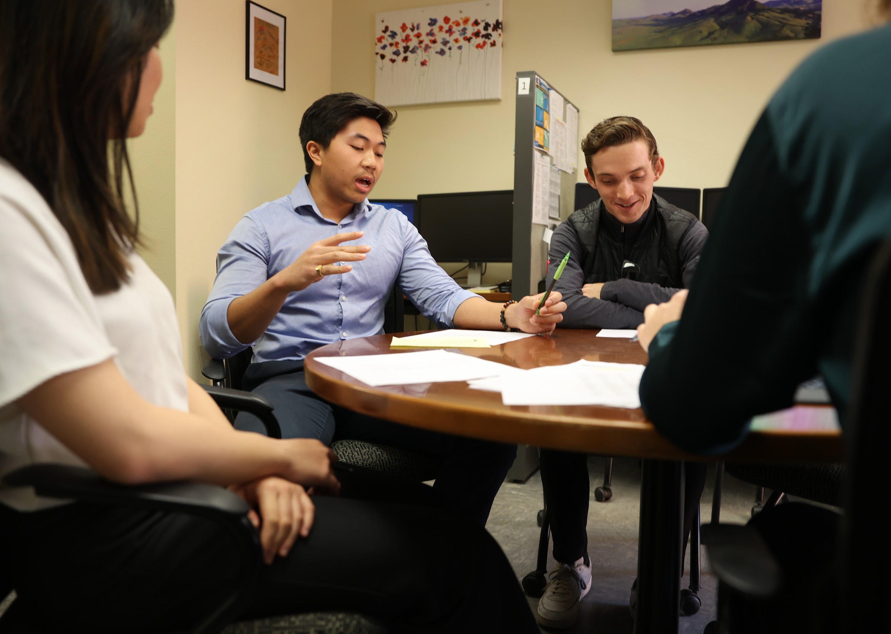 A student in a blue button down speaks to a group of students while gathered around a round table. 