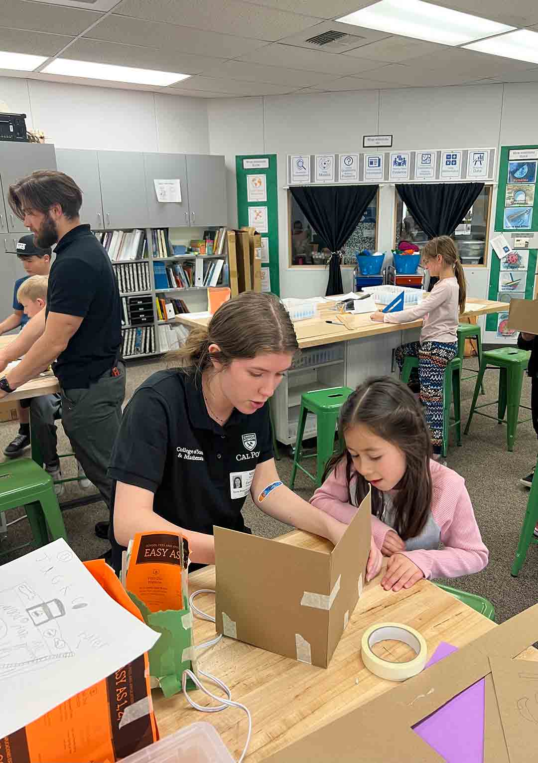 A Cal Poly student in a black shirt sits at a desk and helps a young child with a construction made of cardboard pieces.